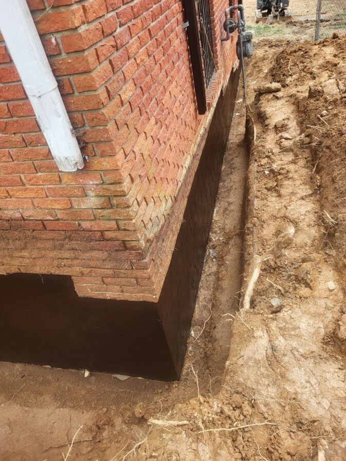 Brick house foundation with dark waterproofing layer applied; trench dug alongside foundation in bare soil.