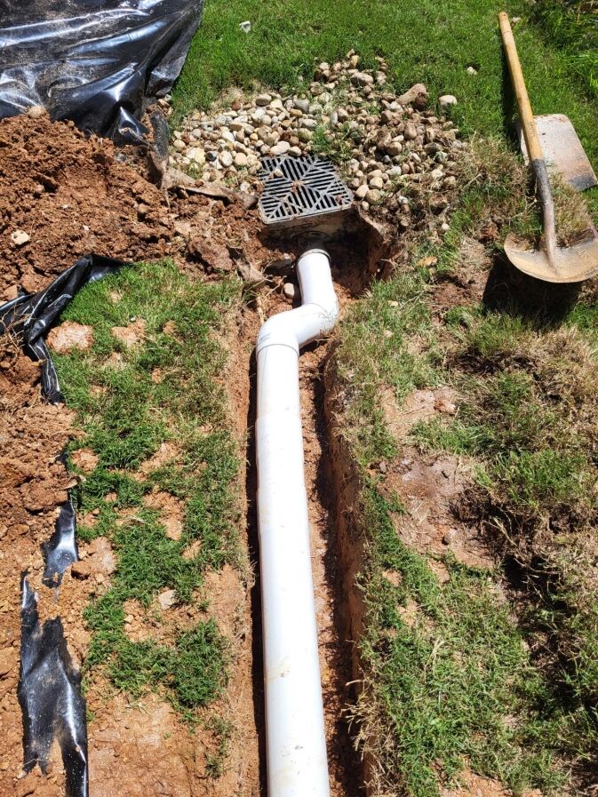 A newly installed white PVC drainage pipe connects to a square grate in a shallow trench with soil, grass, and a shovel nearby.